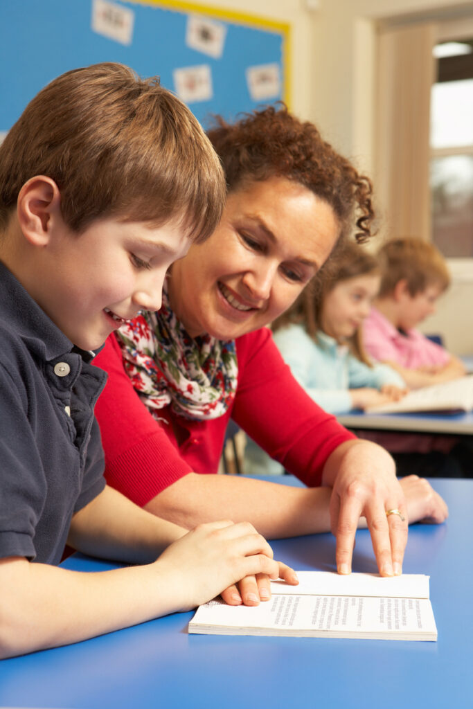 Schoolboy Studying In Classroom With Teacher Reading A Book