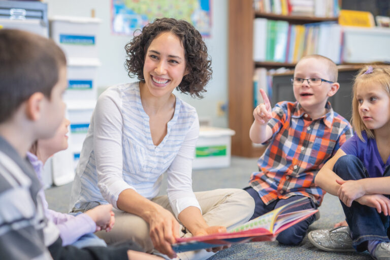 A young ethnic teacher reads a storybook to a group of first graders. They are gathered around her and listening intently as they sit on the floor.